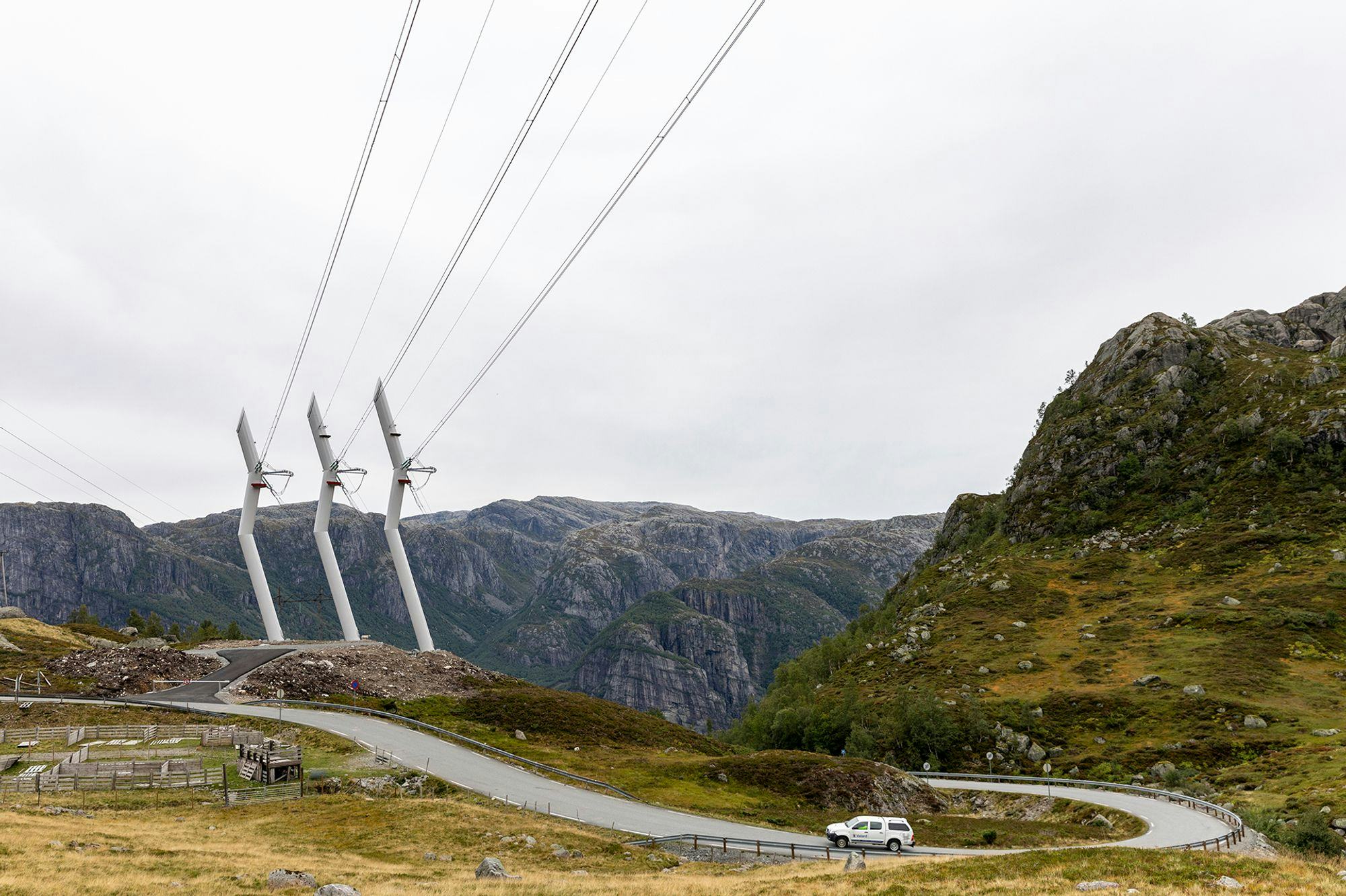 Three single transmission towers in the middle of hills and a white car cruising on a road next to it