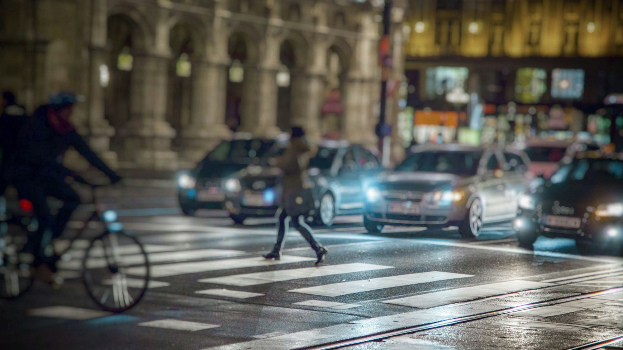 A traffic street in a big city, a crosswalk where a woman walks and a man rides a bicycle. It's dark outside and lights from cars and street lights create a mood