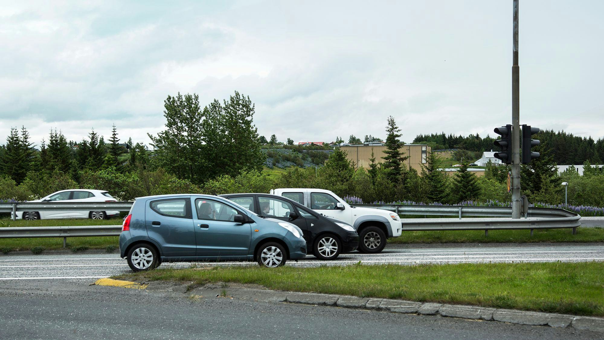 Traffic, three cars driving in the same direction in the middle of the picture, a green area behind the street and a few houses can be seen glimmering